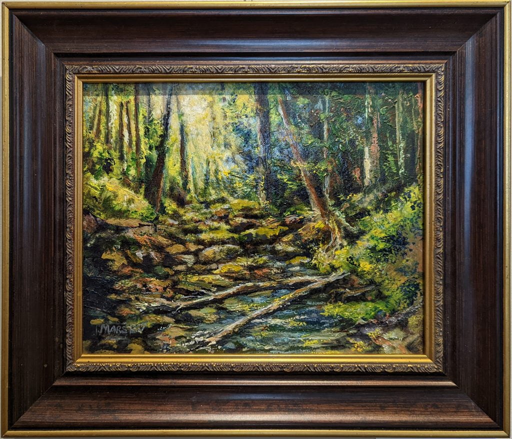 Forest Solitude by Jeanne Marston is an oil painting in a wood and gold-look frame. The painting depicts a very rocky stream running through a forest. The greens and browns, as well as the bright, afternoon light coming through the trees at the top middle-left, give the sense that it is late spring or early summer. The water level is not high, but it is running, and two downed old trees cross the stream at the lower middle. Bright blue sky peeks through the trees on the middle/top right