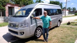 Left side front and side view of the new, silver 15-passenger van, with Carlos Manuel, the cathedral's long-time bus driver, proudly standing beside it, hand on the windowsill,  in jeans, a short sleeved, teal button-down shirt, and sunglasses.