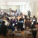 Dr. Jay Fluellen stands at the back of the photo, facing the viewer, behind a keyboard, speaking to a crowd of seated people in the foreground with their backs to the viewer.