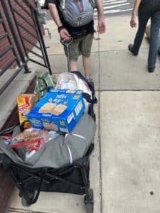 A black wagon, filled of food and other supplies is pulled by a person along a concrete sidewalk. The person is walking away from us. We can only see them from the shoulders down. They are wearing a black t-shirt and dark khaki green shorts and carrying a grey backpack on their back. Another person, walking beside them on the right is also visible, legs down. They are wearing dark pants, t-shirt, and boots. At the left of the photo you can see part of a front stoop to someone's home or business made of brick and concrete and a black railing. At the top of the photo it appears the sidewalk is meeting an intersection with a crosswalk.